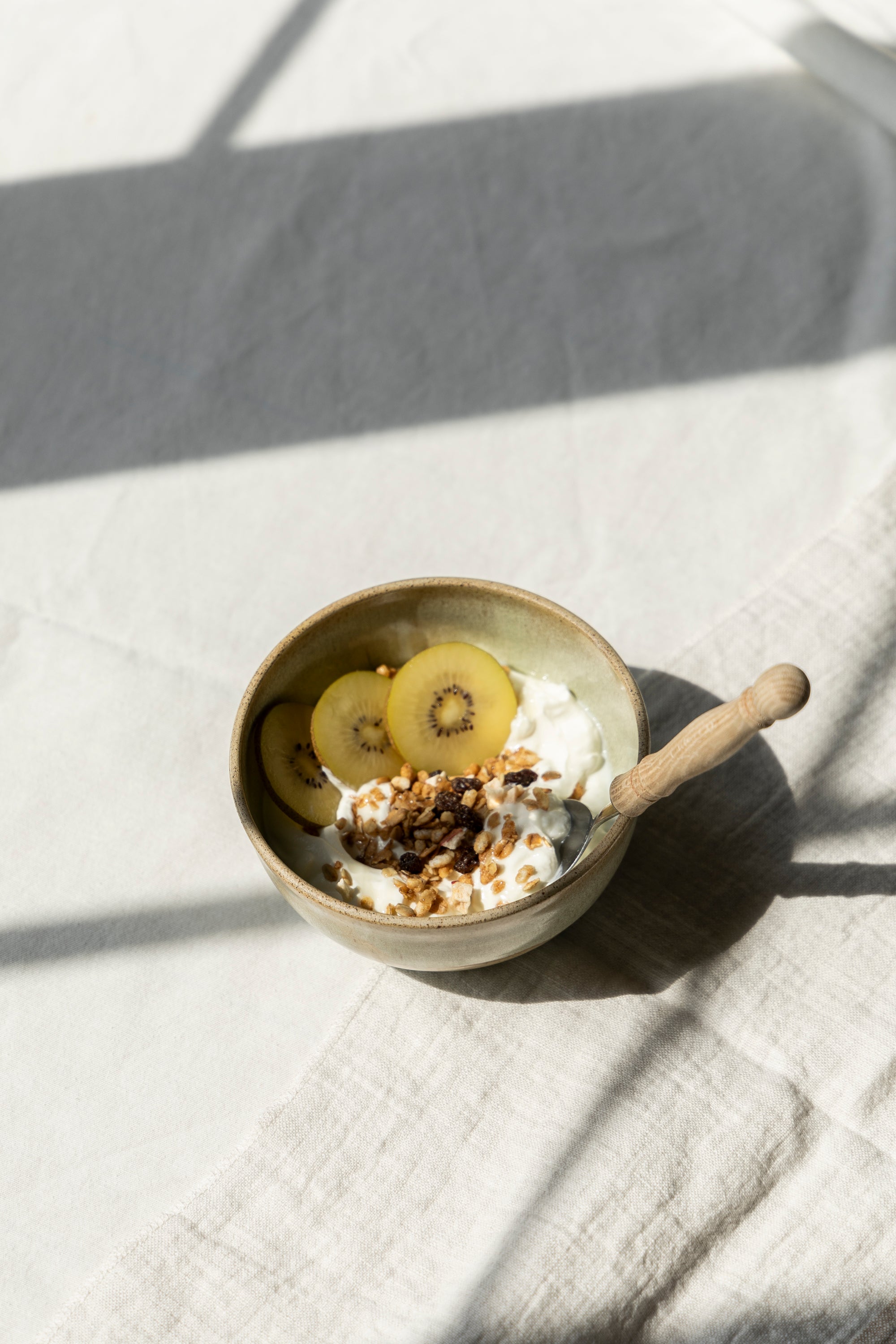 Kom met yoghurt, granola en schijfjes gele kiwi op een linnen tafelkleed in zacht ochtendlicht.